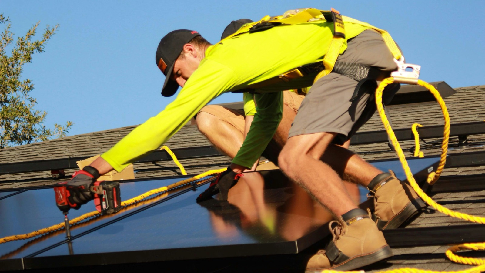 Installer in high-visibility gear securing a solar panel to a shingle roof with an impact drill
